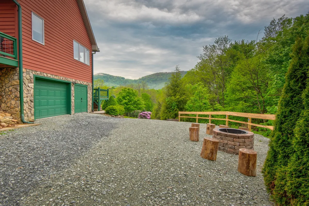 A close-up outdoor setting featuring a stone fire pit with wooden seating, a green garage door, and mountains in the backdrop. The arrangement evokes communal evenings spent around the fire pit, ideal for storytelling and stargazing under open skies.
