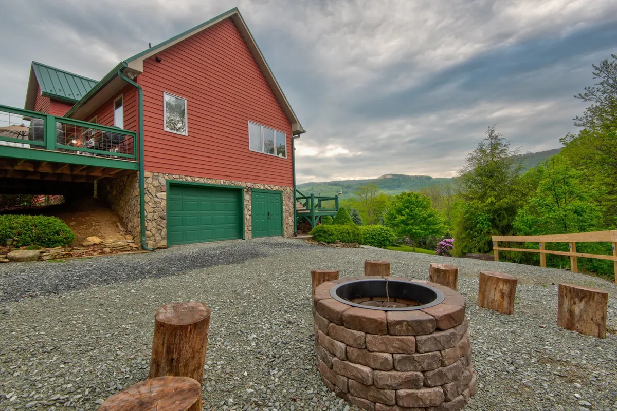 A fire pit area with timber benches, set in front of a red house and surrounded by greenery and mountain views. The outdoor environment invites guests to gather, share stories, and stargaze in comfort.