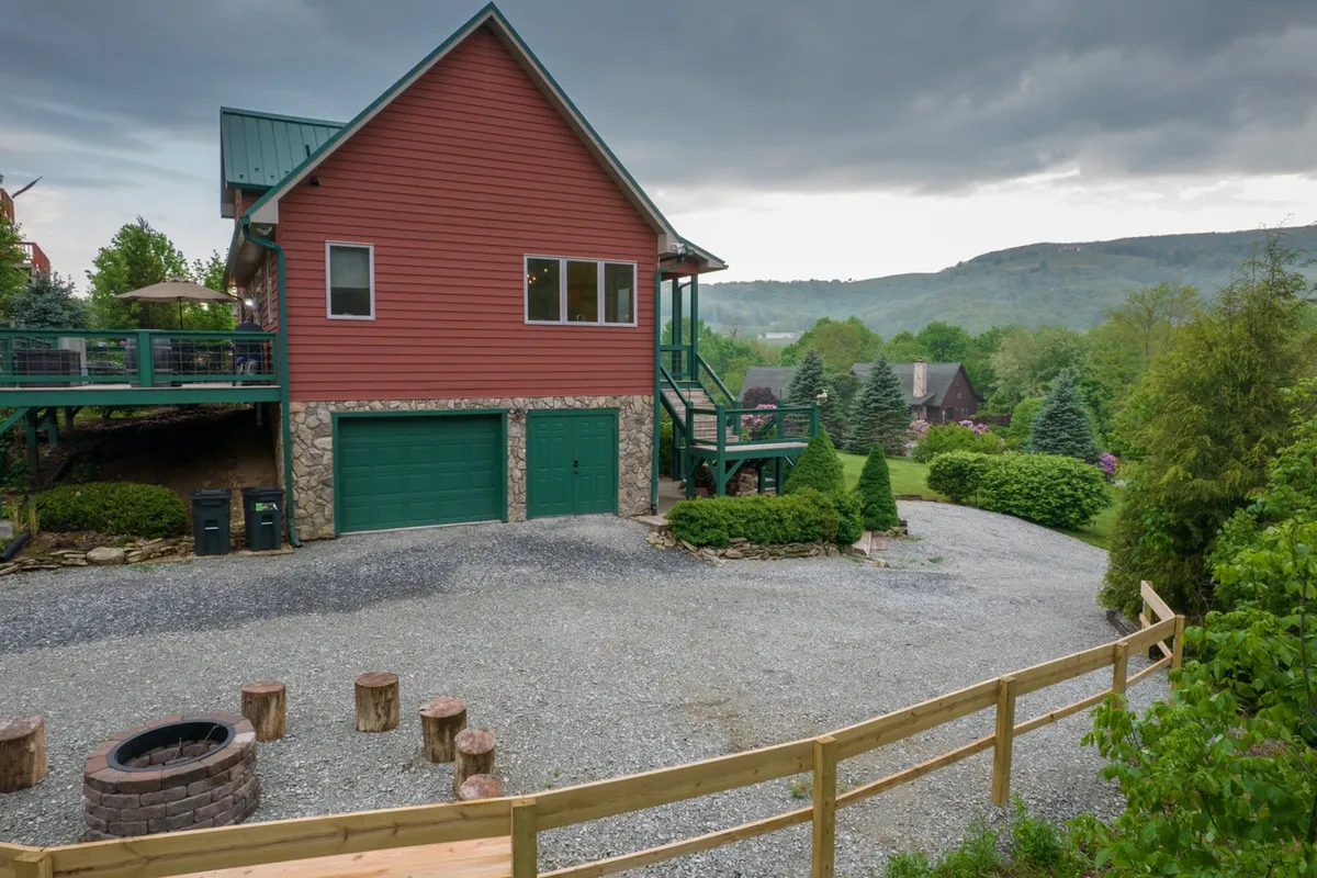 A sweeping exterior view of the red house with a green roof, a welcoming deck, a stone fire pit surrounded by wooden benches, and lush green surroundings. The fire pit area is set against a backdrop of trees, creating a perfect spot for family gatherings and storytelling under the stars.