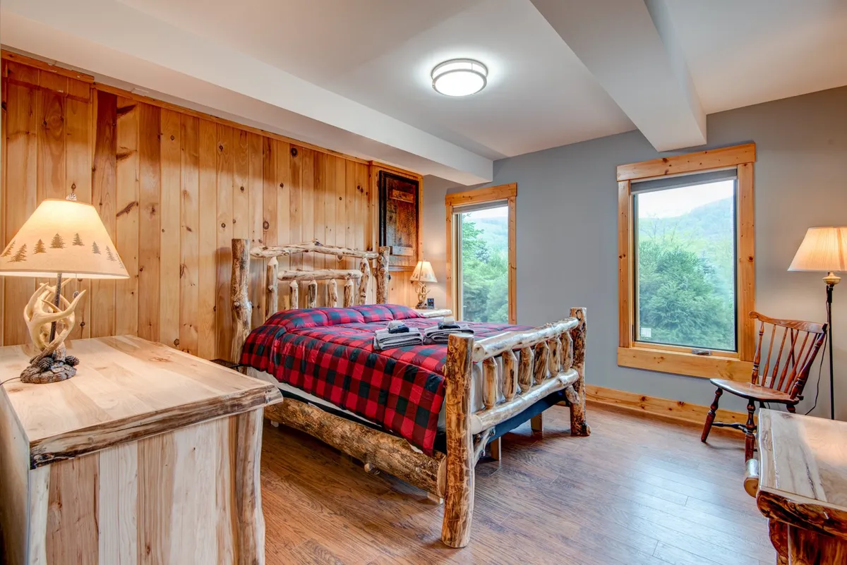 A rustic bedroom showcasing a log bed with plaid bedding, a wooden nightstand, and large windows letting in natural light and offering a view of the outdoors. The inviting reading nook by the window adds a peaceful touch.