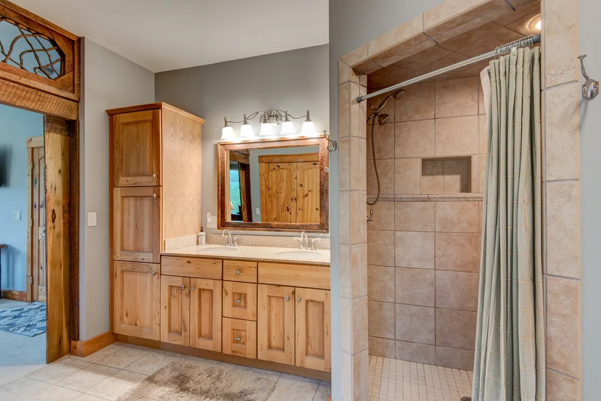 A bright, modern bathroom with a double vanity set in wooden cabinetry, a large mirror illuminated by elegant light fixtures, and a walk-in tiled shower. The space feels both luxurious and practical for shared routines.