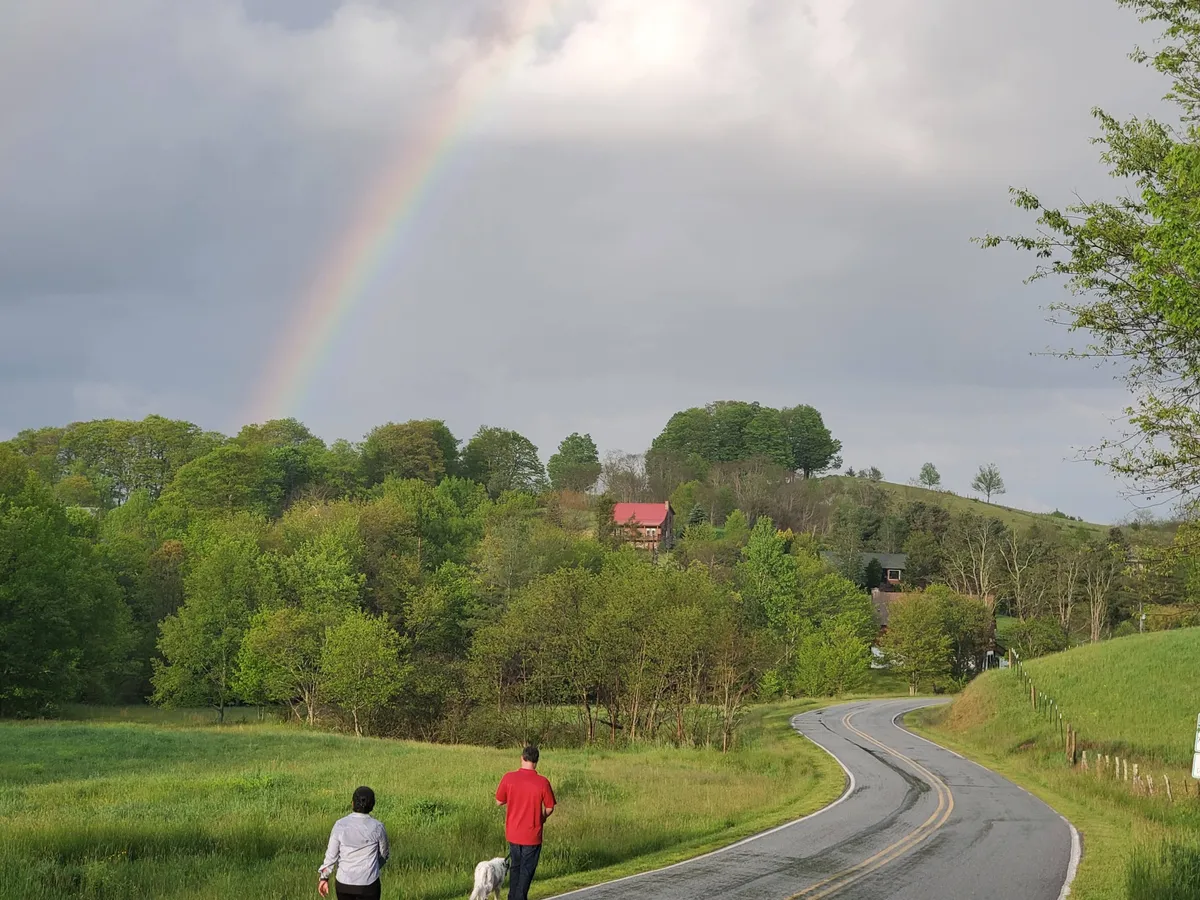 A scenic countryside view featuring a winding road, a vibrant rainbow arcing overhead, people enjoying a walk, and lush greenery stretching into the distance. A distant house is visible, enhancing the sense of open space and exploration.