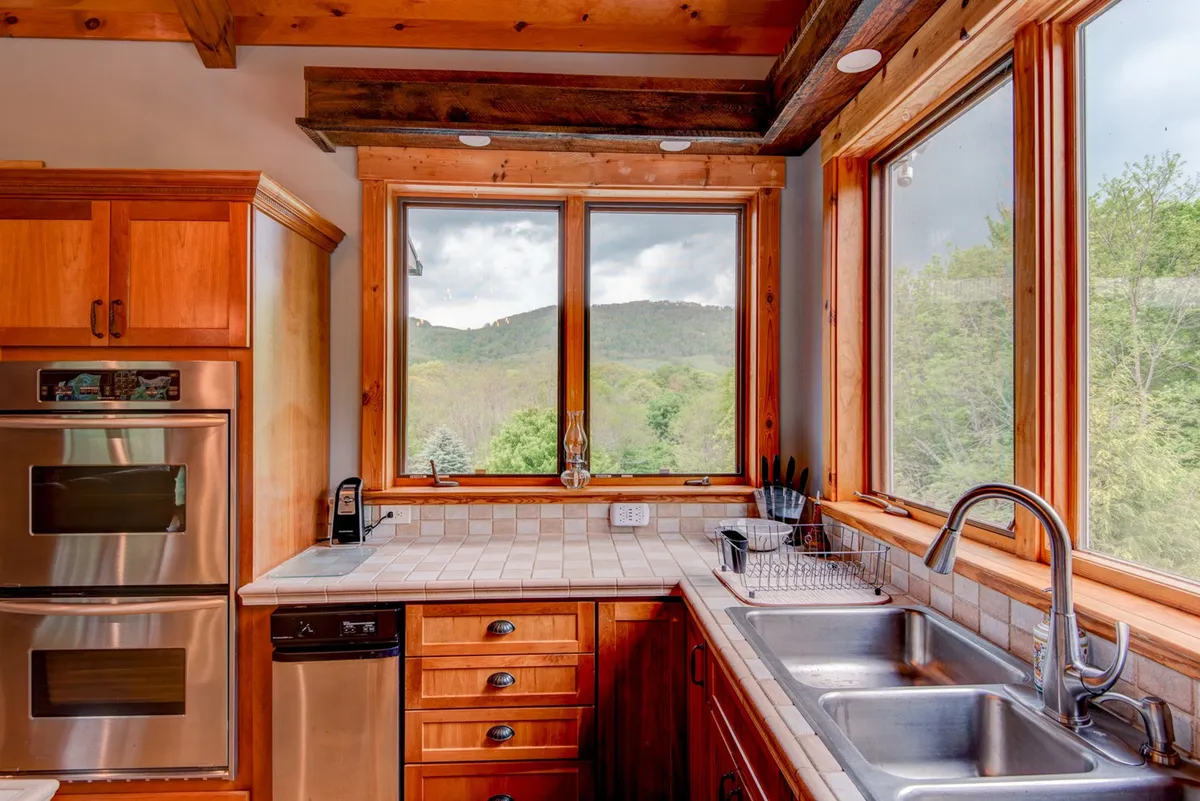 A cozy kitchen corner featuring large windows with breathtaking mountain views, wooden cabinetry, stainless steel appliances, and a ceramic sink. The workspace is designed to maximize both utility and scenic enjoyment, perfect for meal preparation or morning coffee.