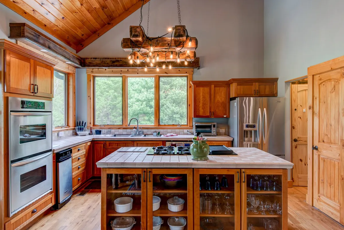 This image captures a warm, inviting kitchen space with a large central island, rustic light fixtures, wooden cabinetry, and expansive windows that reveal lush greenery outside. Stainless steel appliances are visible, and the kitchen appears thoughtfully stocked and ready for both cooking and entertaining.
