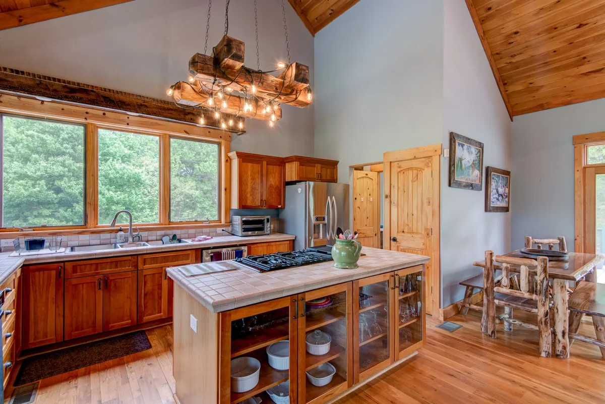 An open kitchen and dining area with large windows looking onto greenery, a rustic chandelier above the table, custom wooden cabinetry, and an island with a glass front. The space feels cozy and communal, ideal for family-style meals and celebrations.