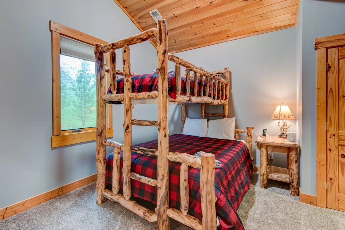 A cozy bunk room featuring a wooden bunk bed with plaid bedding, a wooden nightstand, and a lamp. The space feels inviting and warm, ideal for children or teens to unwind after a day of adventure.