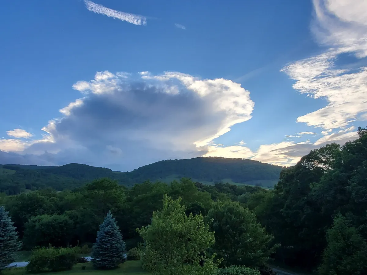 A wide view of rolling hills and lush greenery beneath a dramatic sky filled with clouds. The serene landscape suggests opportunities for nature walks, photography, and relaxation in the outdoors, with hints of sunset lighting.
