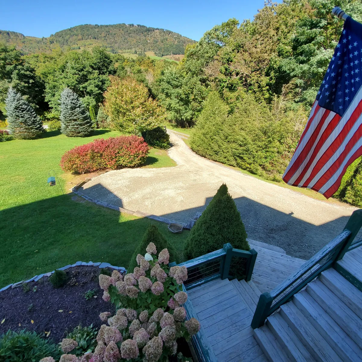 An exterior image highlighting an American flag, flowering bushes, a gravel driveway, and distant mountains. The inviting porch and garden are visible, set against a peaceful mountain backdrop.