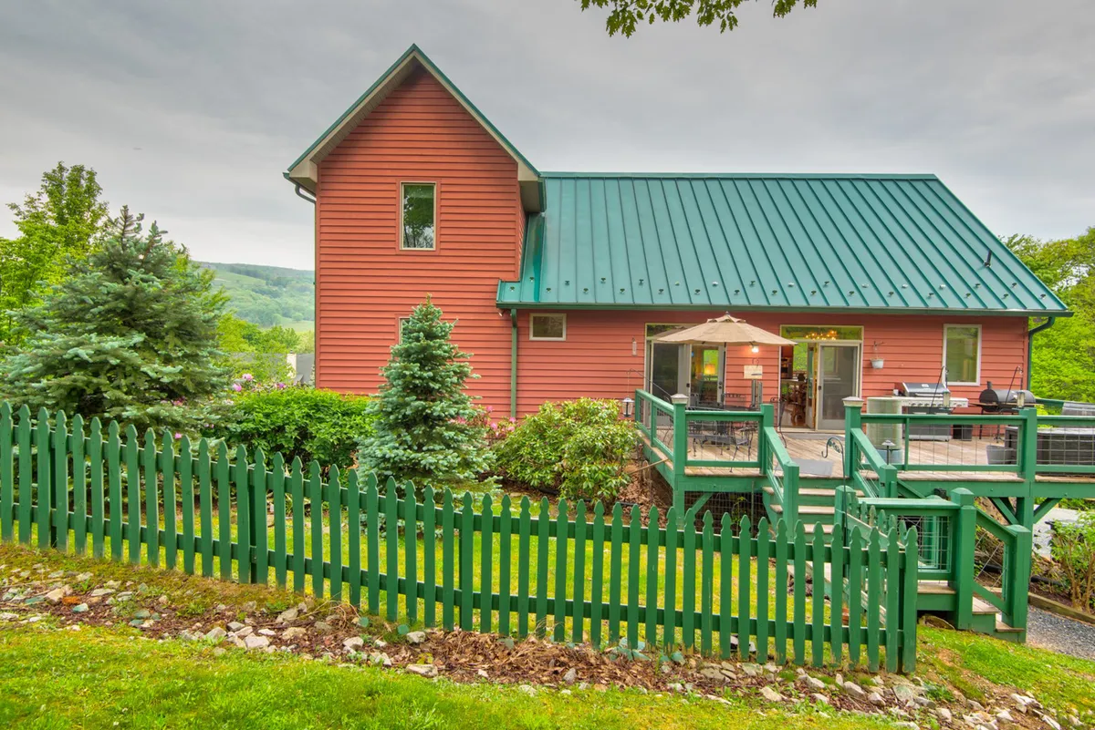 This image features the vibrant pink exterior of the house with a green metal roof, a welcoming wooden deck with outdoor seating, a manicured lawn, and a surrounding fence. The view emphasizes the inviting outdoor living space and the connection to the lush, natural surroundings.