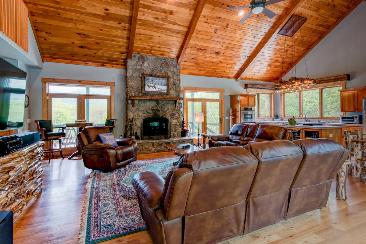A welcoming living room with vaulted wooden ceiling beams, a grand stone fireplace, plush leather sofas, and expansive windows revealing sweeping mountain views. The space is bathed in natural light and arranged for both relaxation and social gatherings.