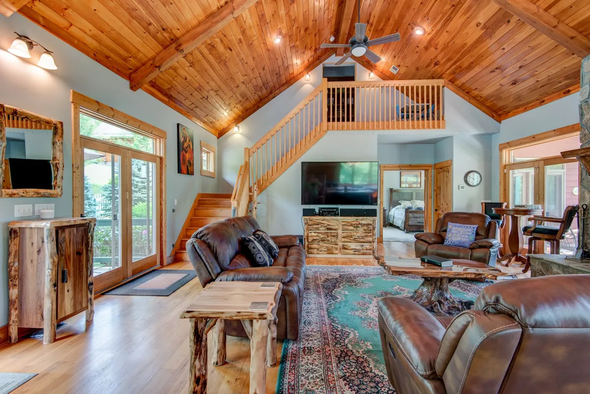 A bright, open living area with exposed wooden beams, a large stone fireplace, comfortable seating, and large windows. The layout encourages socializing, relaxation, and enjoying panoramic vistas outside.