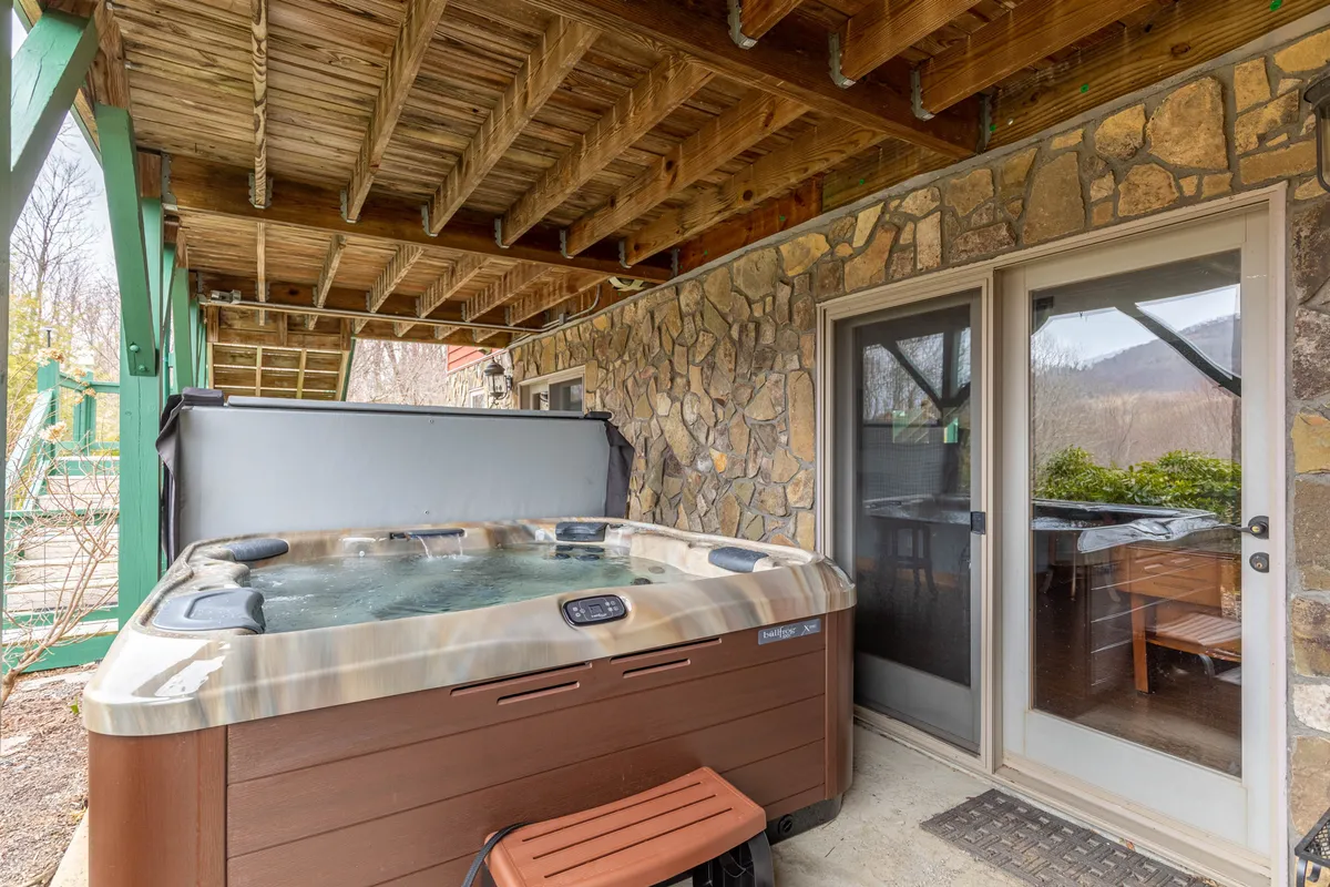 A tranquil indoor-outdoor scene with a hot tub set against a stone wall, large sliding glass doors opening to the outside, and natural light flooding the space. The setup suggests soaking under the stars and seamless integration between relaxation and nature.
