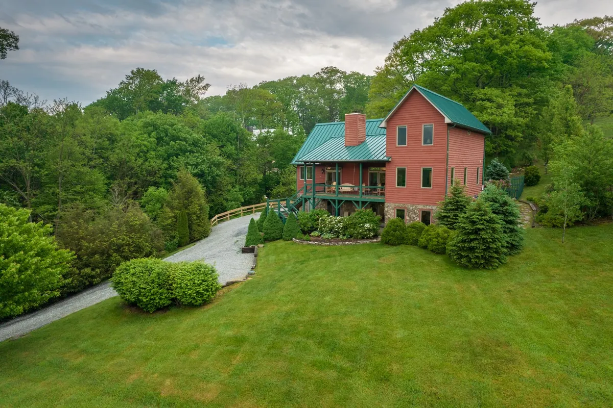 The image showcases a charming farmhouse set against a lush green lawn, surrounded by mature trees and a peaceful countryside backdrop. A gravel pathway leads up to the welcoming front porch, inviting guests to relax and take in the mountain views. The scene exudes warmth, tranquility, and the promise of memorable outdoor gatherings.