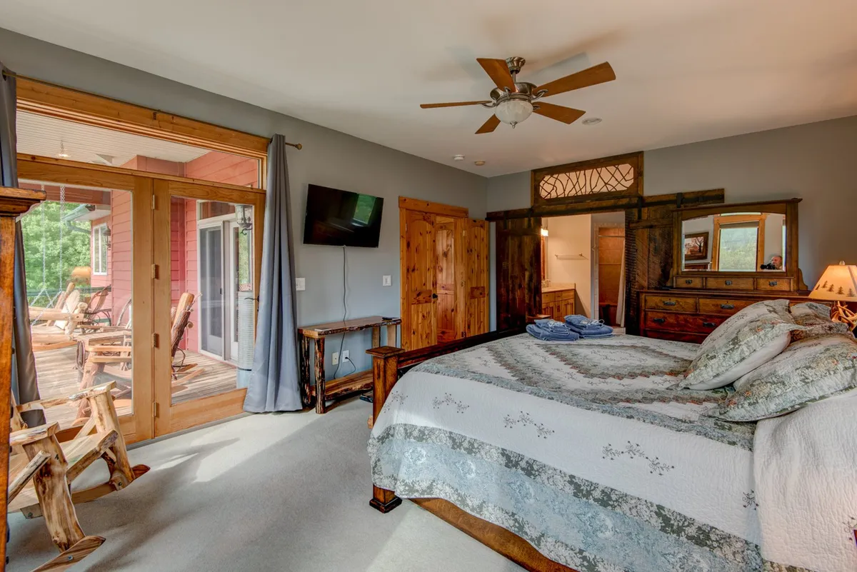 A serene bedroom with a king-sized bed dressed in patterned bedding, complemented by rustic wooden furniture and sliding glass doors that open onto a deck overlooking nature. The soft natural light and inviting decor create a calming atmosphere, ideal for restful mornings and evenings.