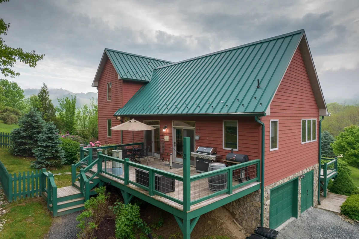A welcoming exterior shot showcasing the farmhouse’s deck furnished with patio furniture, set against a backdrop of lush greenery and the home's distinctive red wooden siding and green metal roof. The spacious deck hints at ample room for gatherings, al fresco dining, and relaxation in nature.